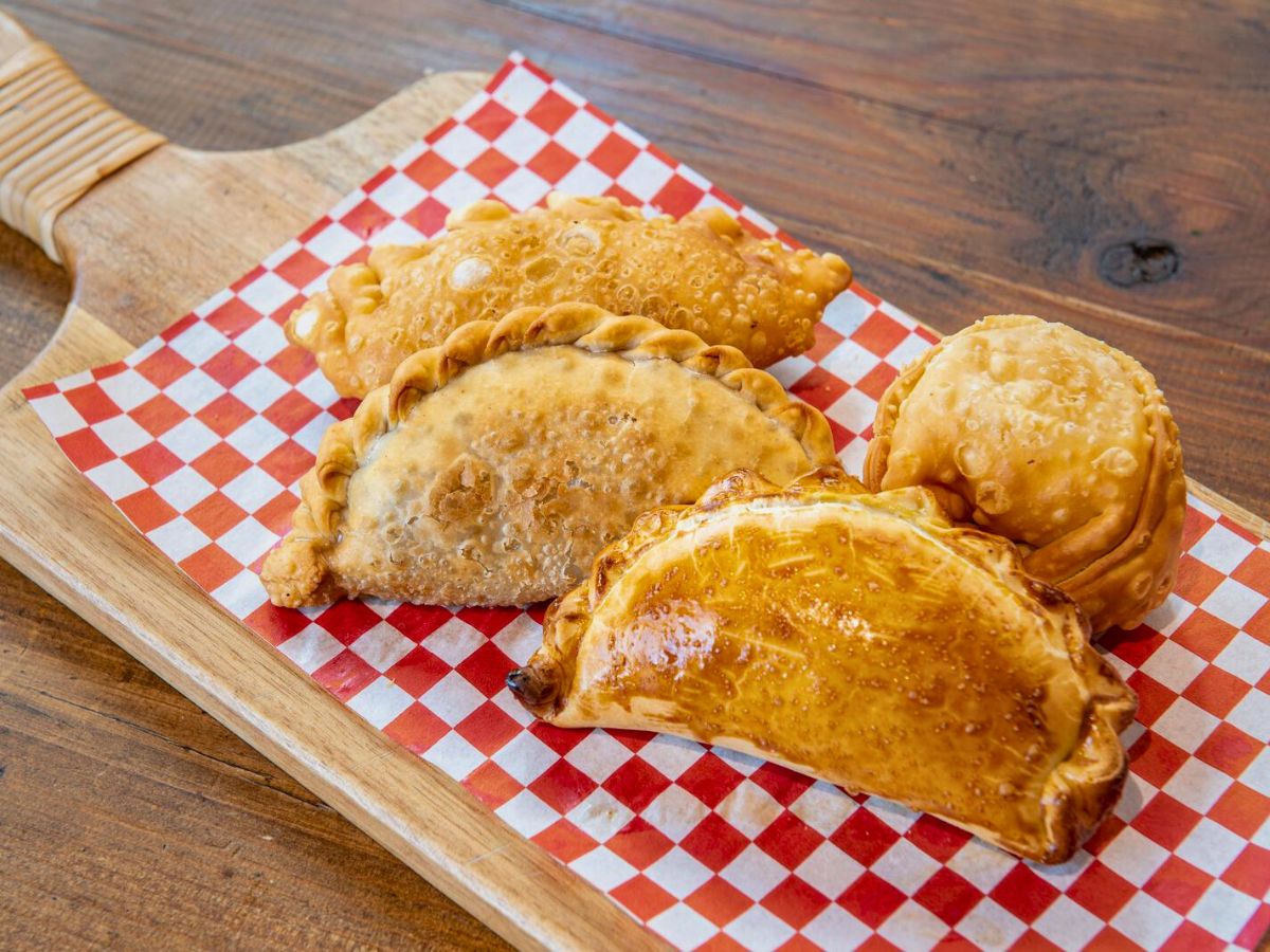A wooden board with a red and white checkered paper holds four assorted empanadas, each with different fillings, placed on a wooden table.