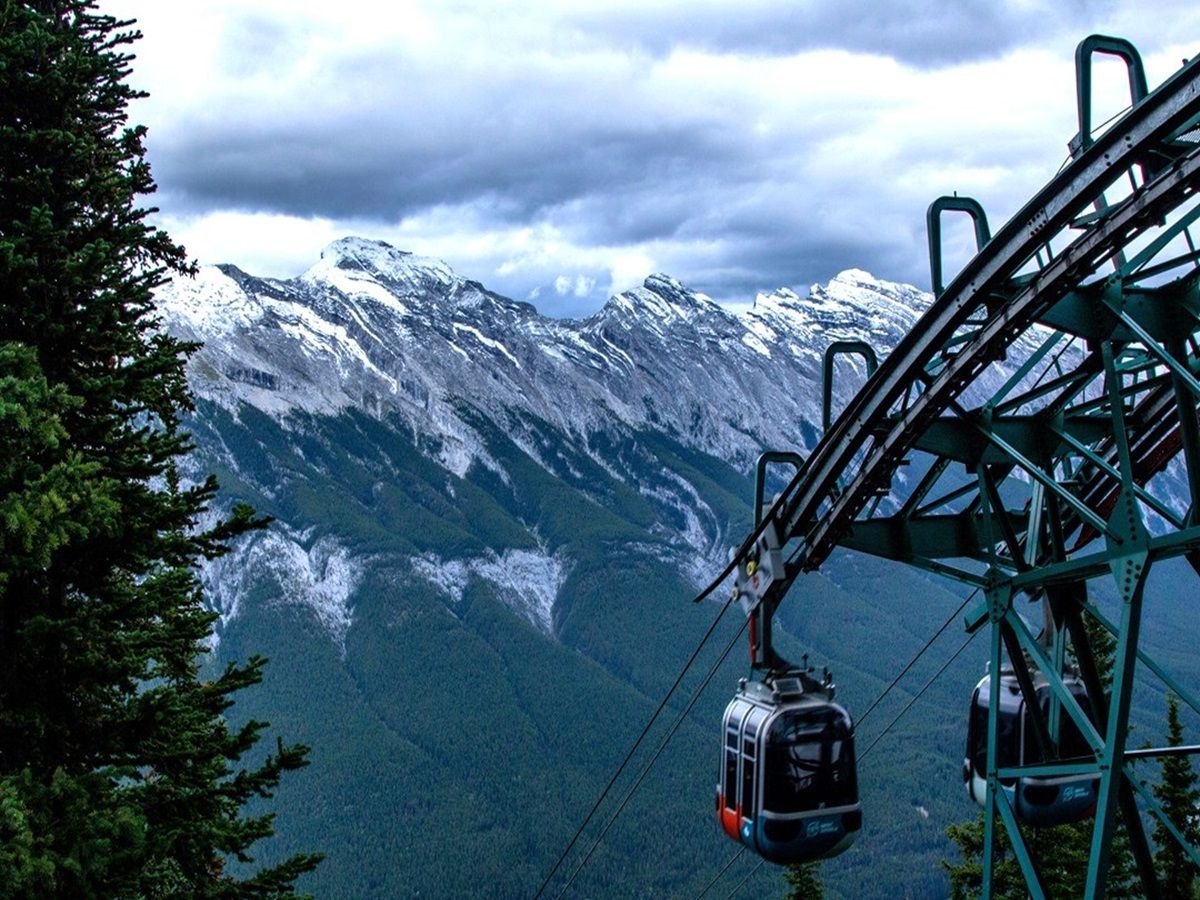 Cable cars ascend a mountain against a backdrop of snow-covered peaks and dense forest under a cloudy sky.