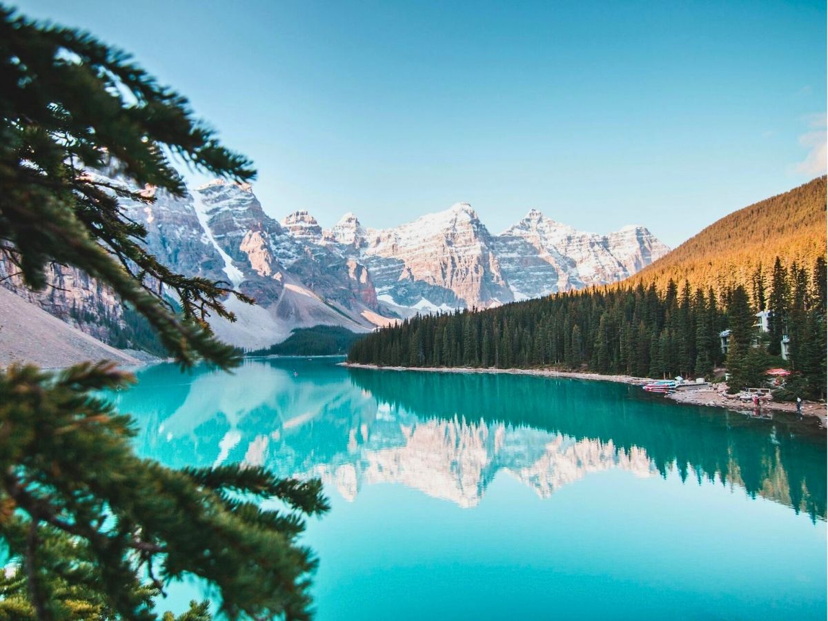 A clear turquoise lake is framed by pine trees, reflecting snow-capped mountains and a bright blue sky, creating a serene natural landscape.