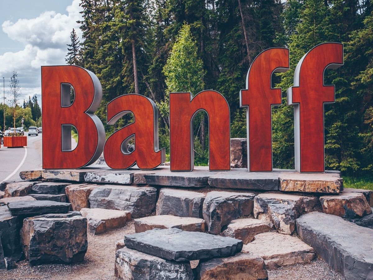 Large red "Banff" sign standing on a rock platform with trees and a cloudy sky in the background.