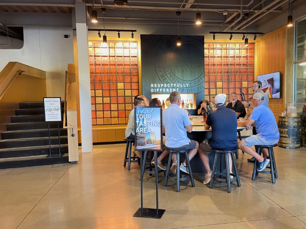 A group of people sit at a round table in a tour tasting area inside a distillery, with a large sign on the wall behind them.