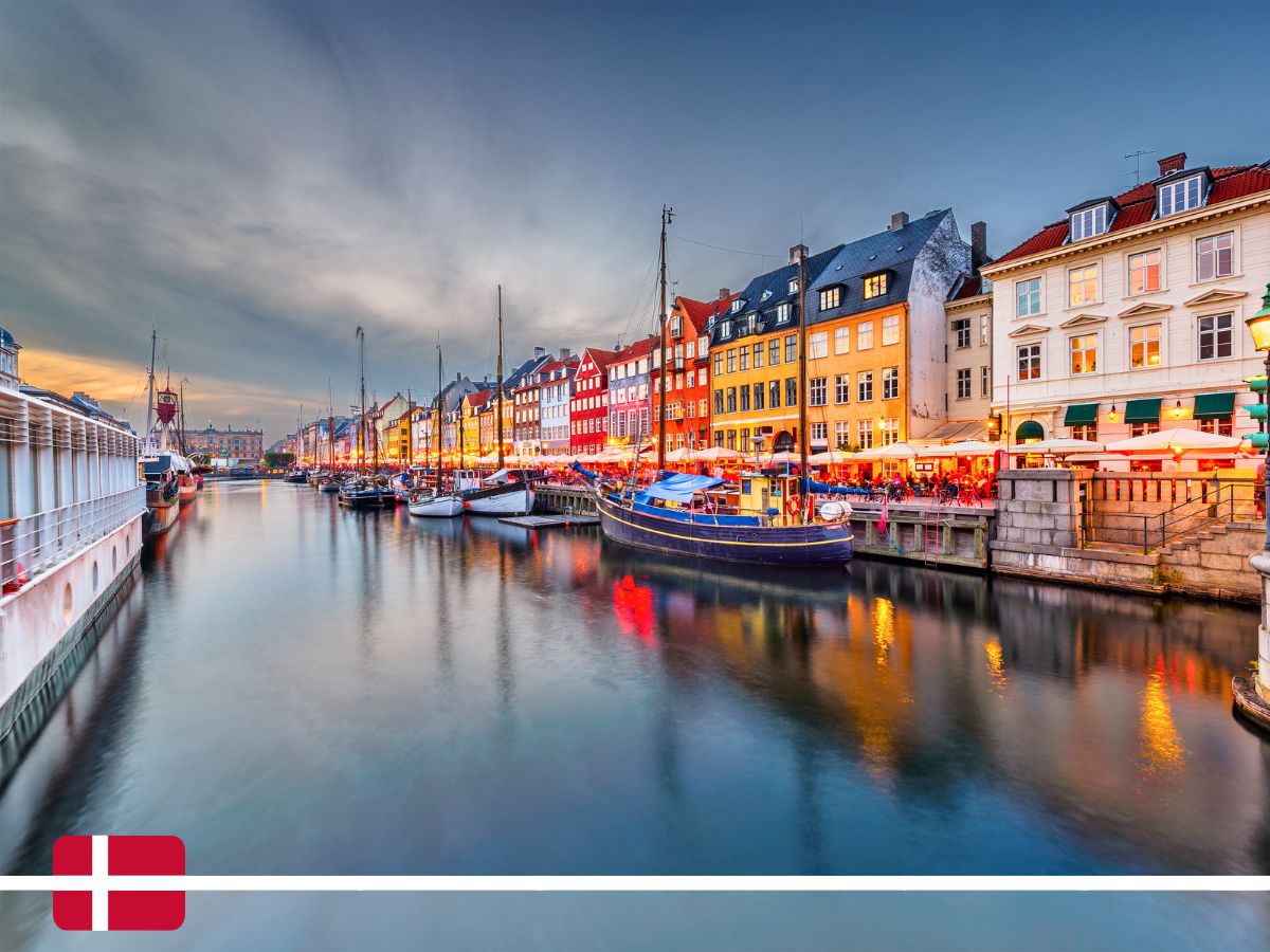 Colorful buildings and boats line the Nyhavn canal in Copenhagen, Denmark, illuminated at dusk with vibrant reflections on the water. A small Danish flag is at the bottom left corner.