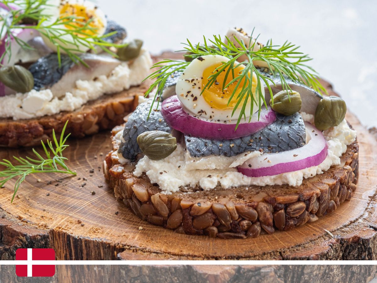 An open-faced sandwich with herring, boiled egg slices, red onion, capers, and dill on a slice of seeded bread with a Danish flag in the corner.