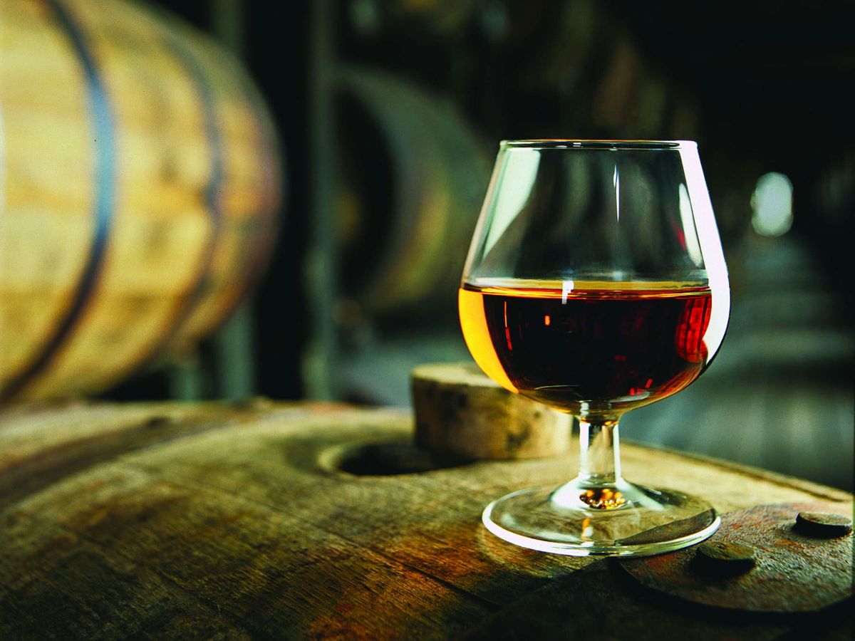 A glass of Kentucky bourbon rests on a wooden barrel in a dimly lit room, with other barrels blurred in the background.