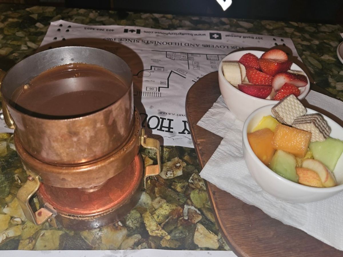 A metal pot filled with chocolate fondue is placed alongside two bowls containing sliced fruits and wafers on a table.