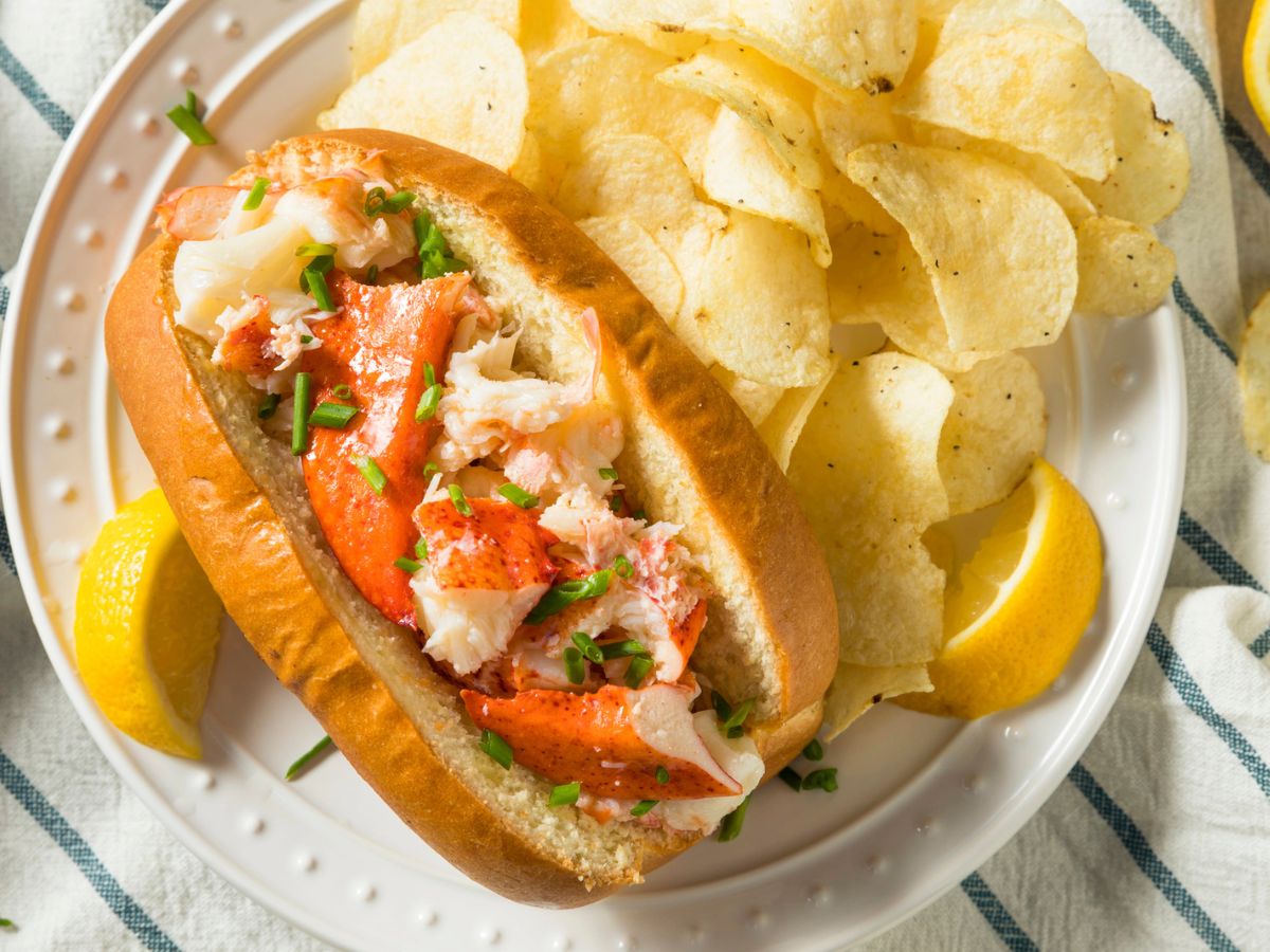 A lobster roll garnished with green chives is served in a bun next to a pile of potato chips on a white plate with lemon wedges.