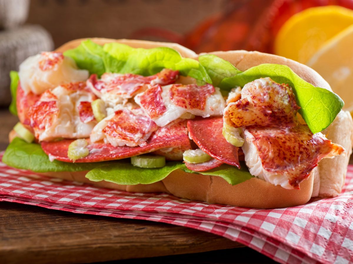 A lobster roll with lettuce on a bun is placed on a red checkered napkin with lemon slices in the background.