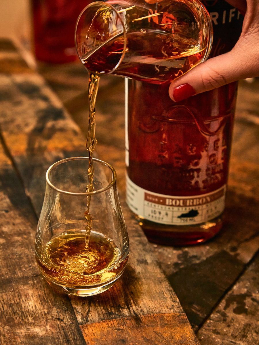 A person pours New Riff bourbon from a bottle into a glass on a rustic wooden surface.