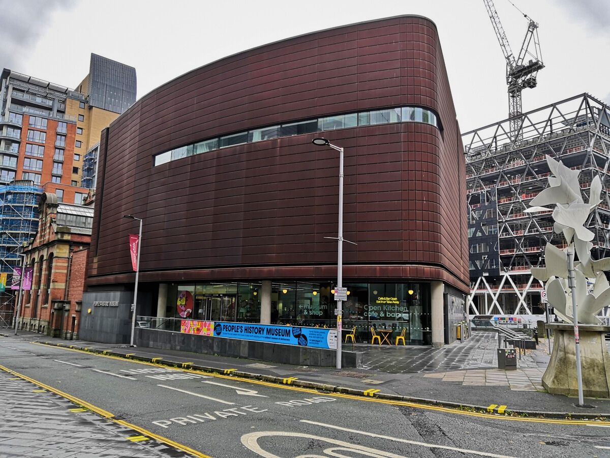 A modern, brown building with rounded corners stands on a city street. Visible signage includes "People's History Museum" and "Exhibition." Construction is seen in the background.