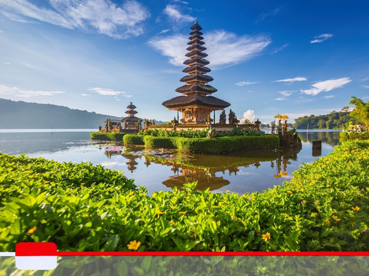 A temple surrounded by water and lush greenery under a clear blue sky, with the Indonesian flag in the bottom left corner.