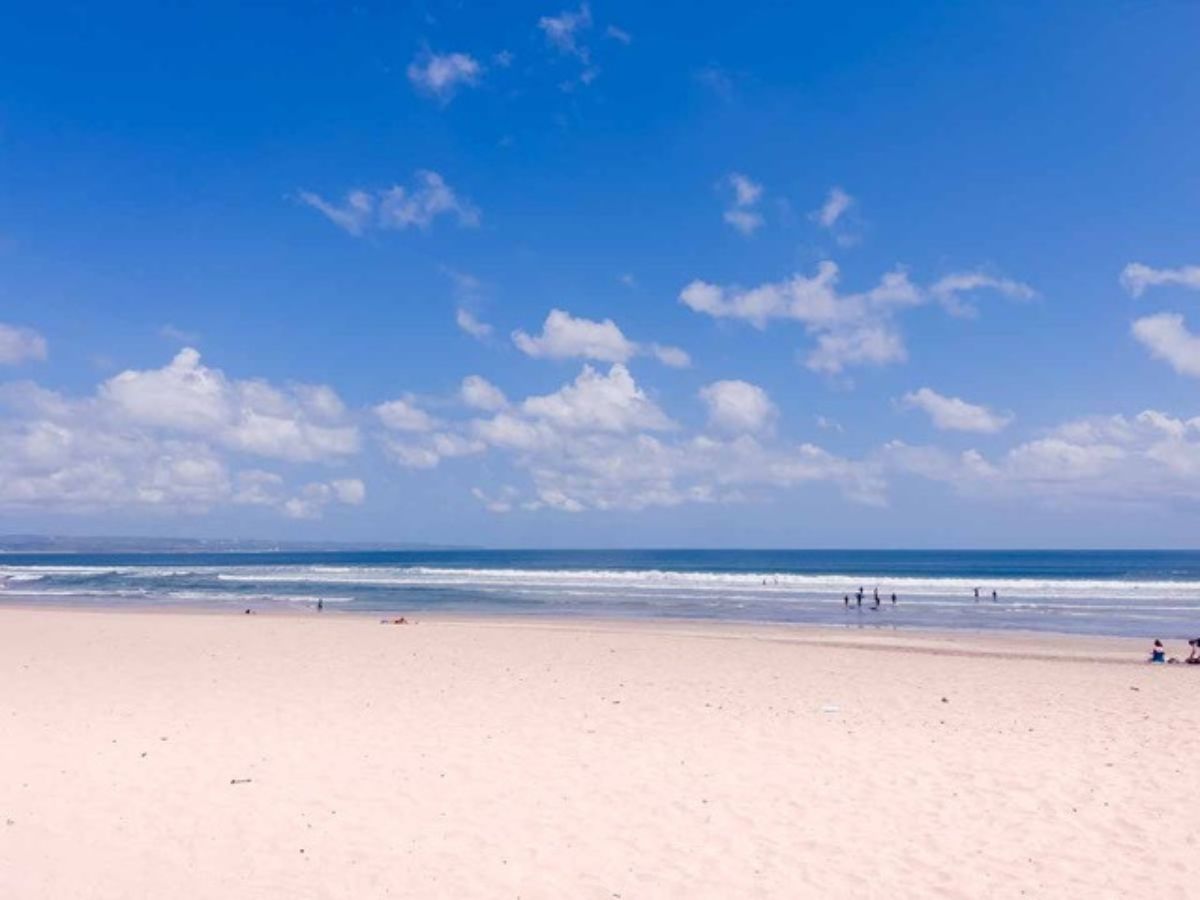 Sandy beach with a few people under a bright blue sky and scattered clouds. The ocean waves are visible in the background.