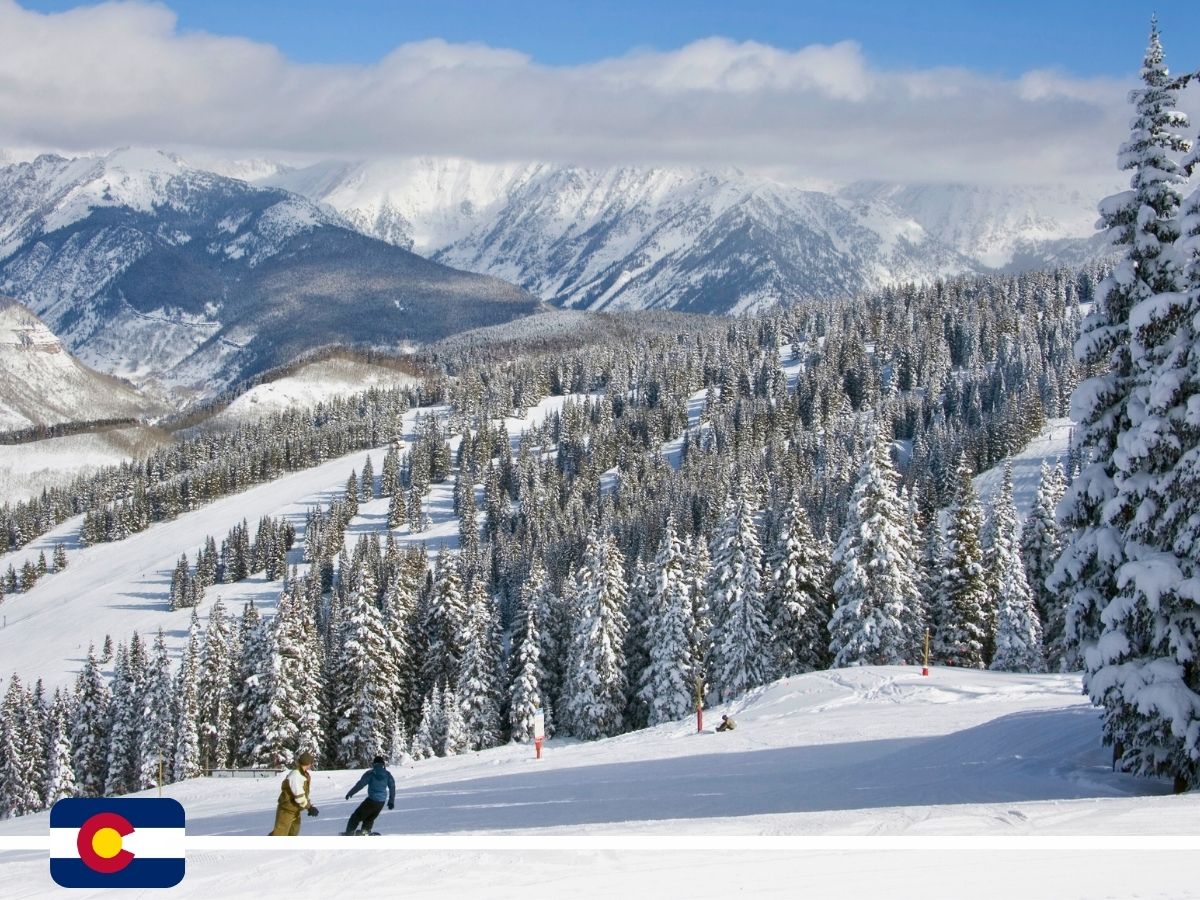 Two people snowboard on a snowy slope surrounded by snow-covered trees and mountains. A Colorado state flag is in the bottom left corner.