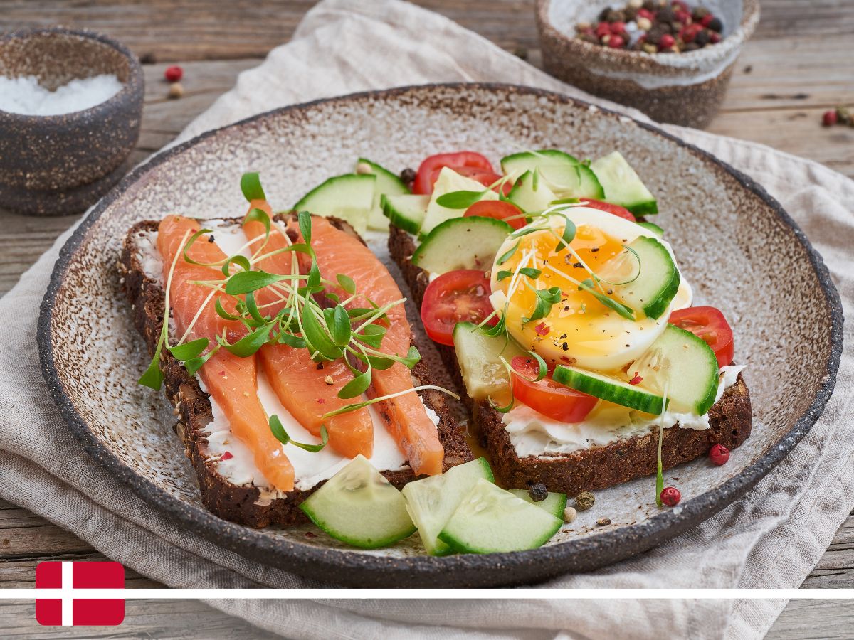 A plate with two open-faced smorresbrod sandwiches on rye bread, one topped with smoked salmon and the other with a poached egg, cucumber slices, cherry tomatoes, and microgreens.