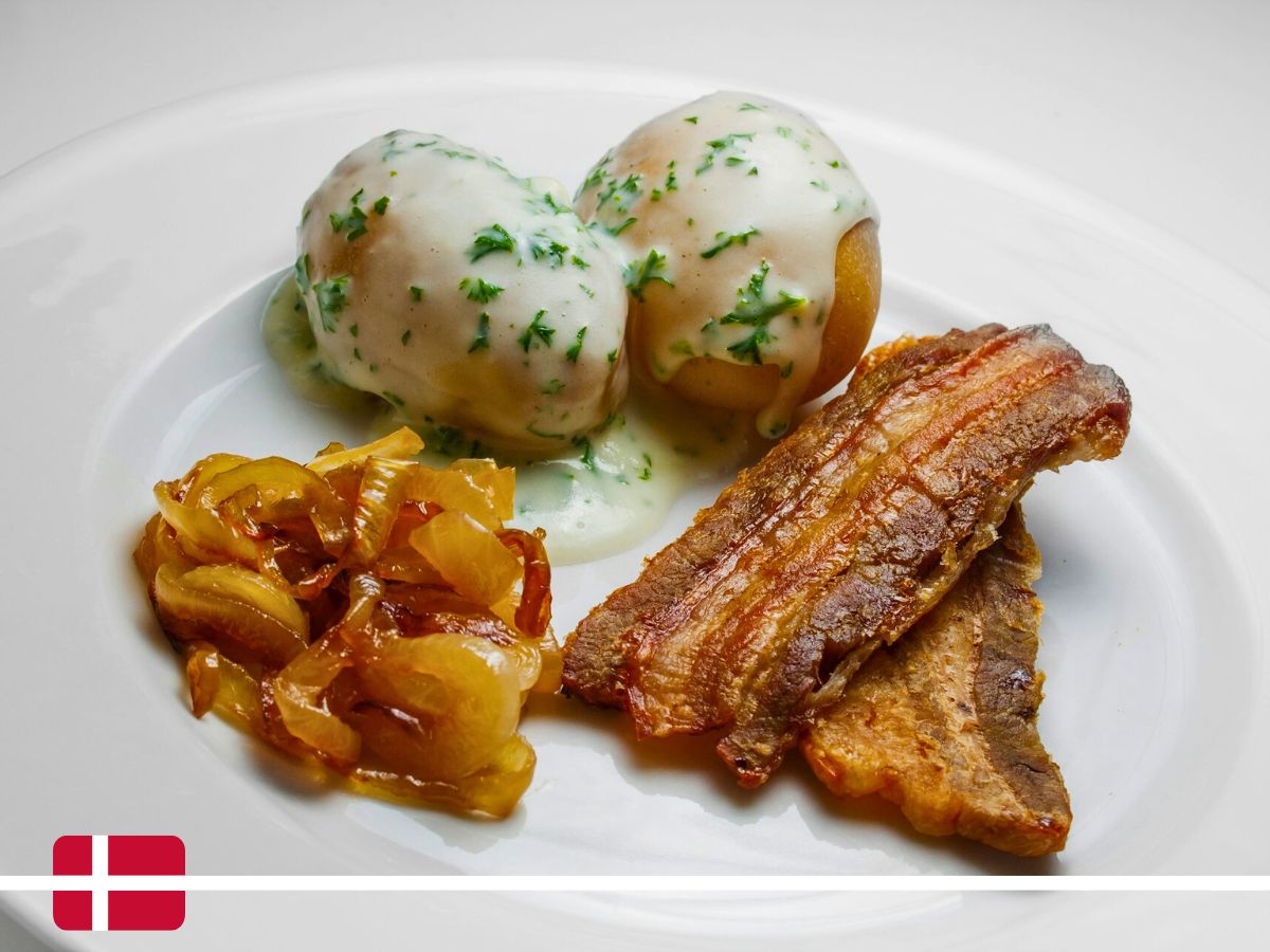 A plate featuring two potatoes with white sauce and herbs, crispy bacon strips, and caramelized onions. A small Danish flag is displayed at the bottom left corner.