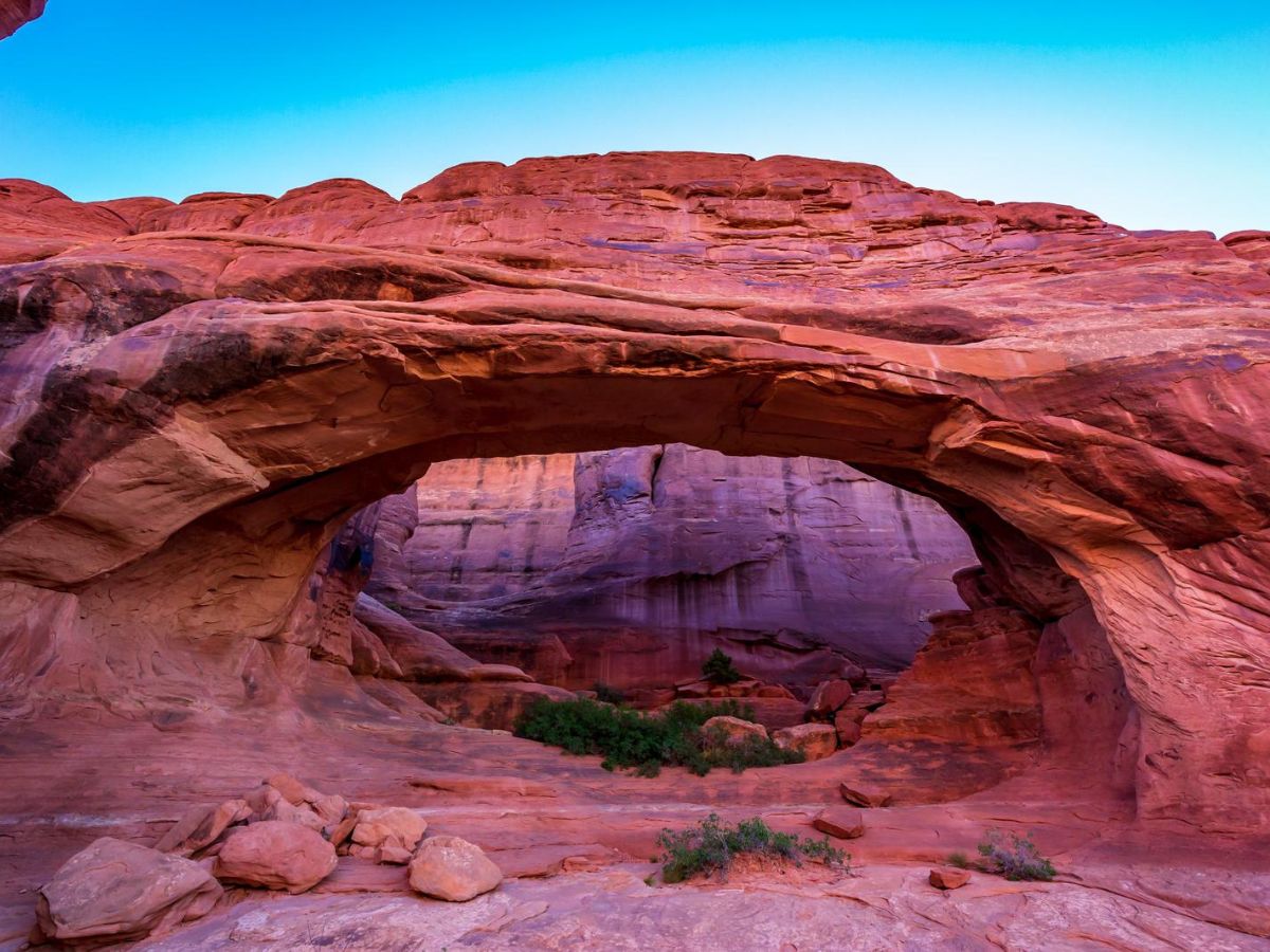 A large, natural red rock arch set against a clear blue sky, with rugged terrain and some green vegetation visible beneath and around the arch.