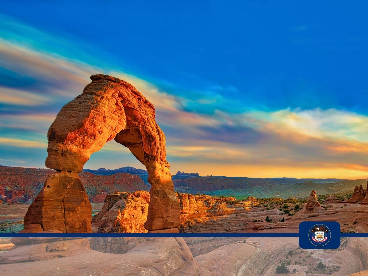 A natural sandstone arch formation is set against a vibrant sunset sky, located in a desert landscape. The Utah state emblem appears in the bottom right corner.