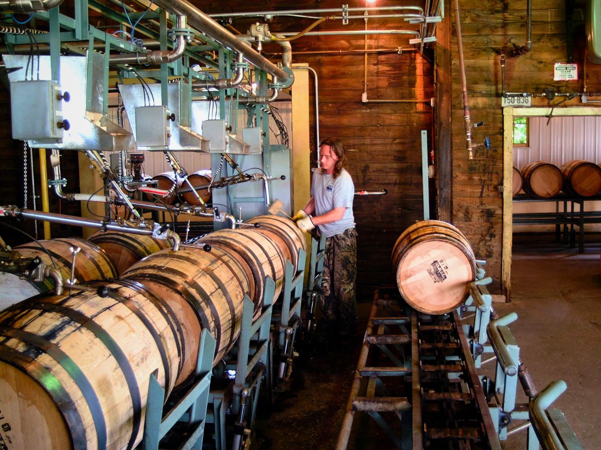 A person operates machinery for filling wooden barrels with liquid in a distillery. Several barrels are lined up and some are already filled.