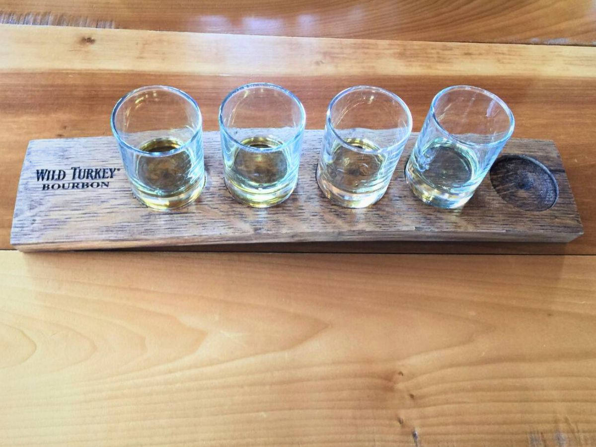 A wooden serving plank labeled "Wild Turkey Bourbon" holds four partially-filled whiskey glasses, arranged linearly, on a wooden table.