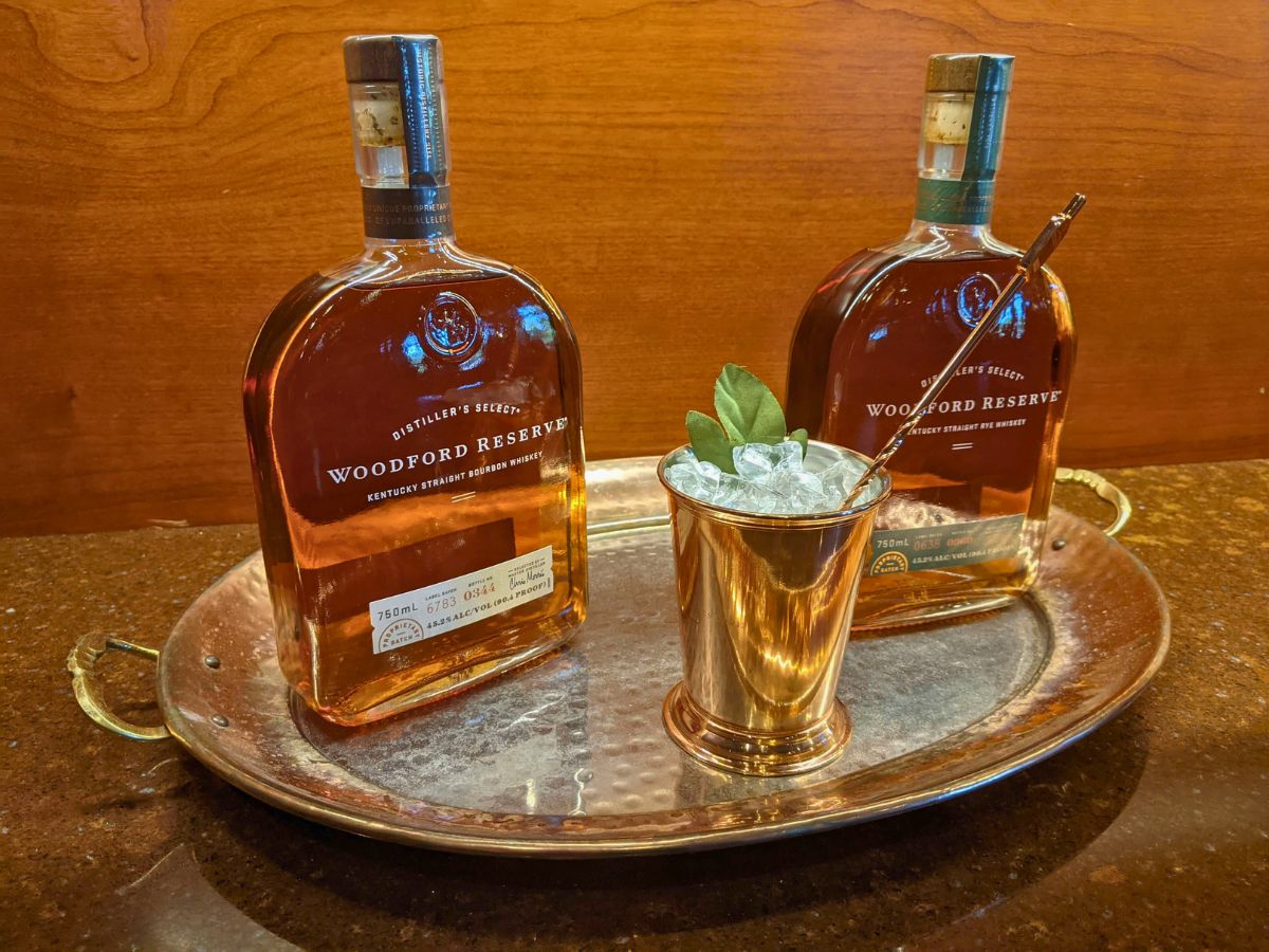Two bottles of Woodford Reserve bourbon sit on a metal tray alongside a copper cup filled with a mint garnish and crushed ice, positioned on a wooden surface.
