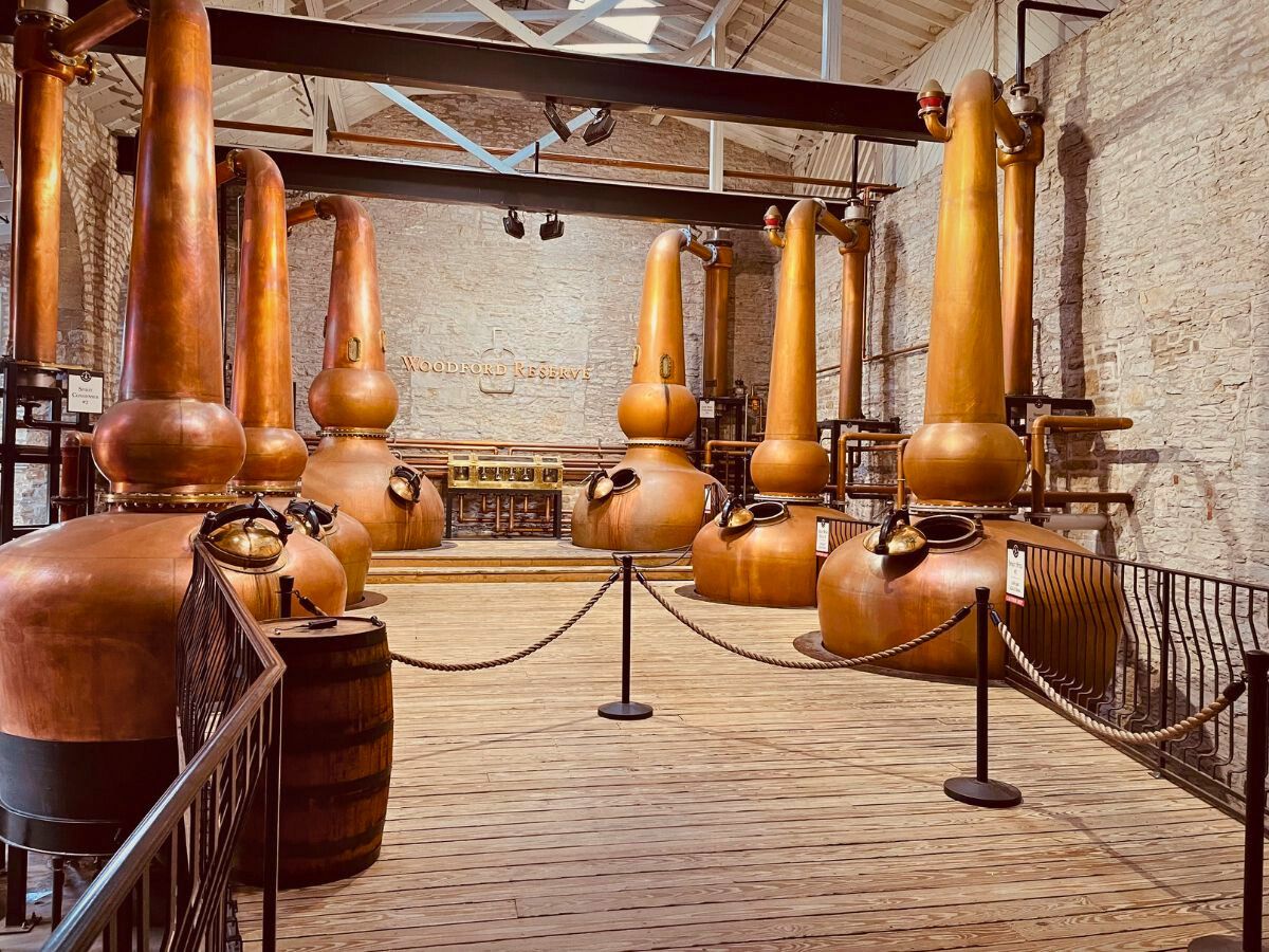 Copper stills and equipment inside the Woodford Reserve Distillery with wooden floors and brick walls, separated by a rope barrier.
