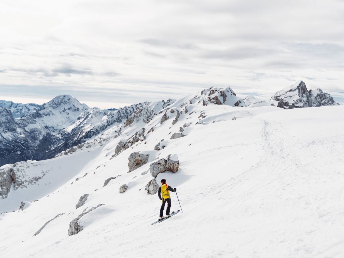 A person wearing a yellow jacket is skiing on a snowy mountain slope with rocky outcrops, surrounded by a mountainous landscape under a cloudy sky.