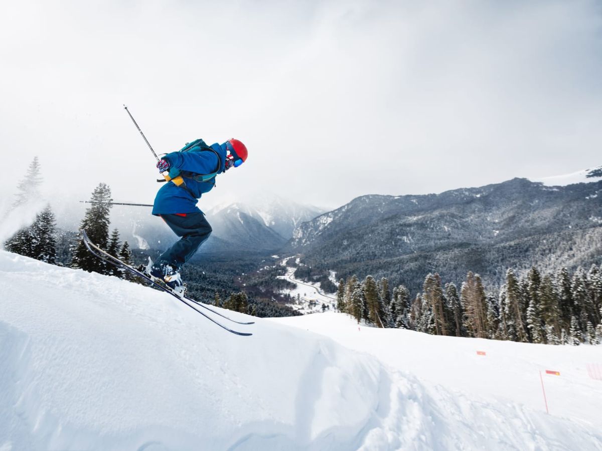 A skier in blue gear and a red helmet is captured mid-air while jumping off a snowy slope with a mountainous landscape in the background.
