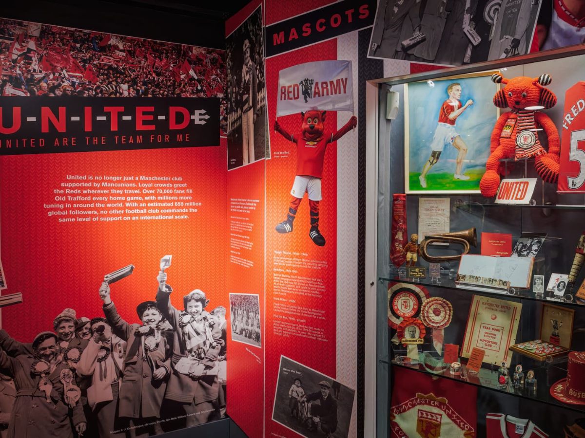 Exhibit on Manchester United's mascots and history, featuring trophies, memorabilia, and a costume of the mascot, Fred the Red. Walls are adorned with photos and descriptions of the club's legacy.