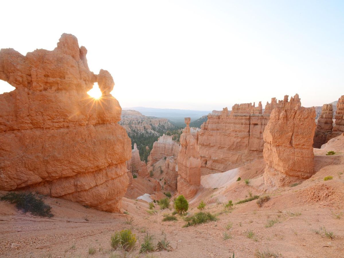A rocky landscape at sunset features orange sandstone formations and hoodoos with a sunburst effect visible through an arch in the foreground. Sparse vegetation covers the ground.