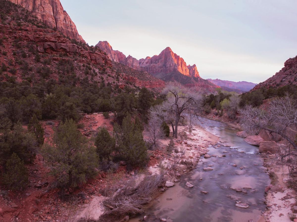 A scenic view of a river flowing through a valley surrounded by rocky, red cliffs and green vegetation under a clear sky during sunset.