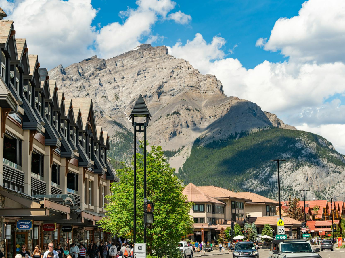 A bustling town with people on the street, cars, shops, and buildings, set against a backdrop of a large mountain range under a partly cloudy sky.