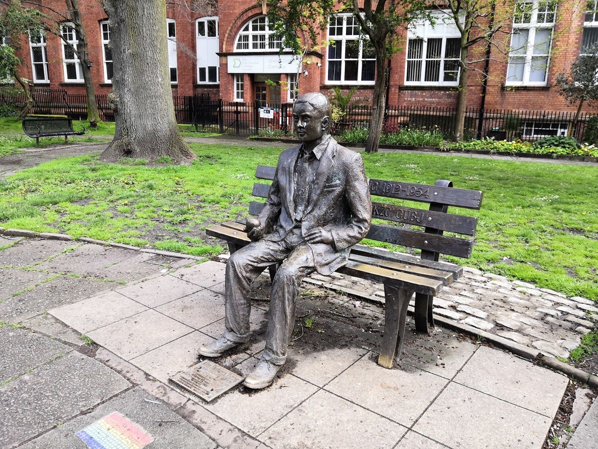 A bronze statue of a man in a suit seated on a wooden bench on a paved area with grass and trees in the background.