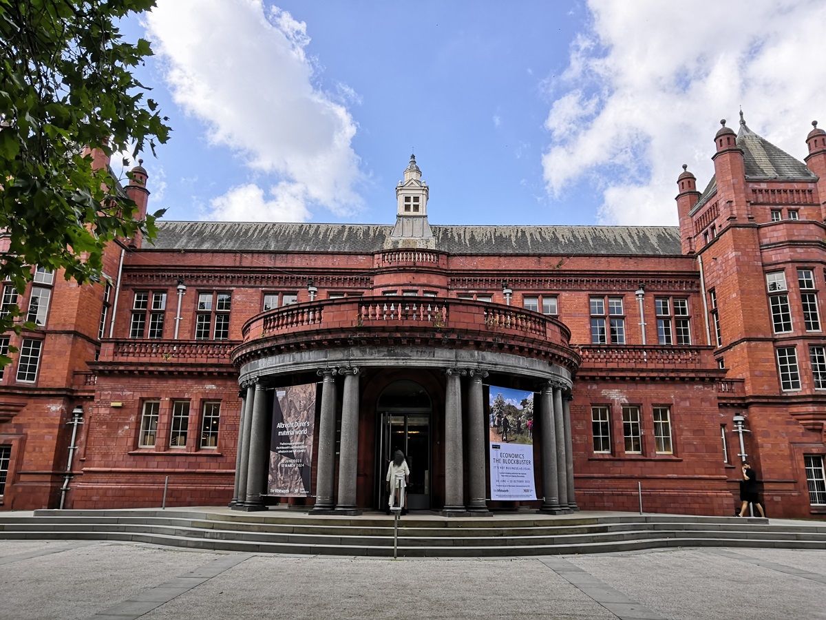 A historic red-brick building with columns at the entrance, featuring banners on either side, under a partly cloudy sky.