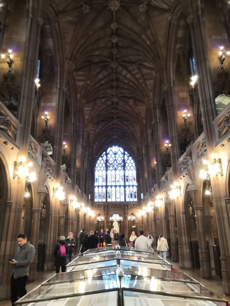 People walk and look at exhibits in a grand, Gothic-style hall with a high, ornate ceiling and a large stained glass window.