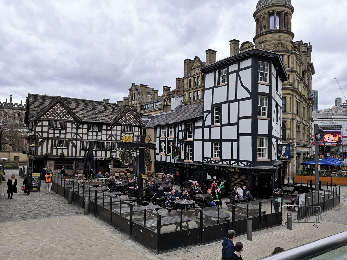 A photo of an outdoor plaza featuring traditional half-timbered buildings with people sitting at outdoor tables; the sky is overcast, and some modern buildings are visible in the background.