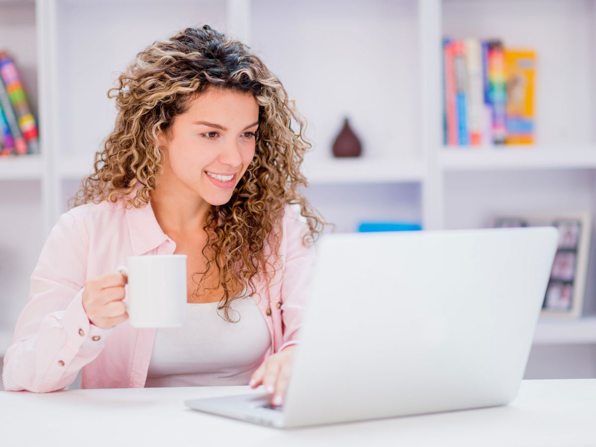 Person with curly hair holding a mug and using a laptop in a bright room with bookshelves in the background.