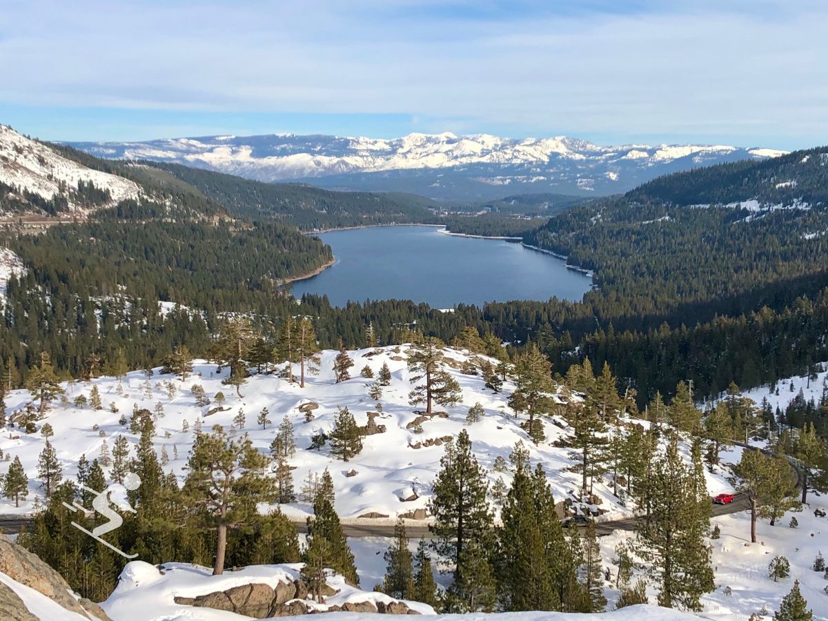 Snowy mountainous landscape with a lake surrounded by pine trees. A winding road cuts through the scene, and distant snow-capped peaks are visible under a clear blue sky.