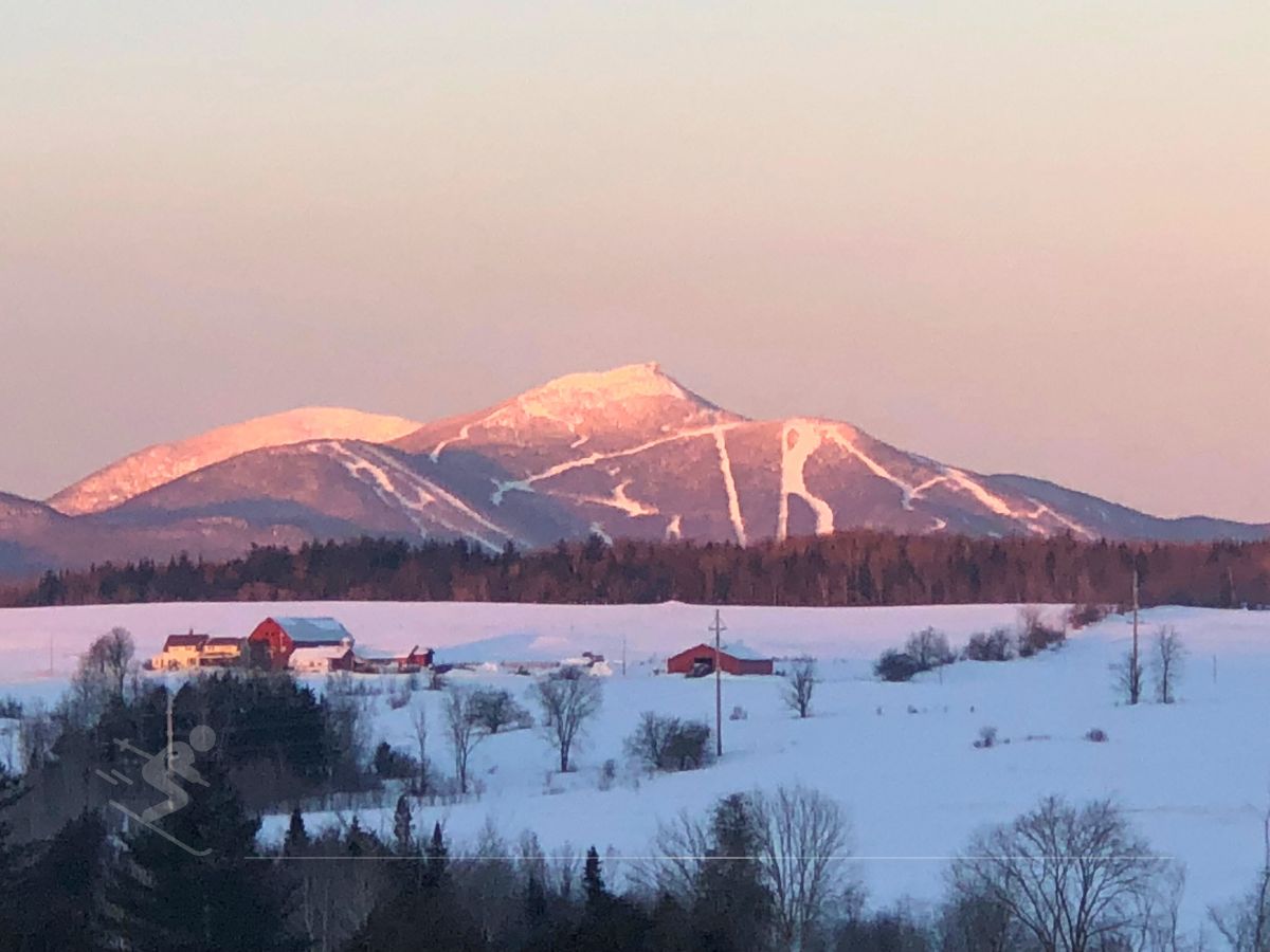 Snow-covered mountain peaks at sunset, ski trails visible. Foreground features a snowy landscape with scattered buildings and trees.