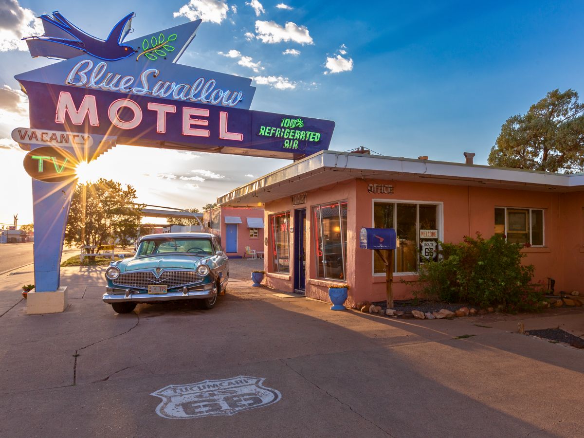 Vintage motel with neon sign reading "Blue Swallow Motel" and classic car parked in front. Sun setting in the background. Signs indicate vacancy and Route 66.