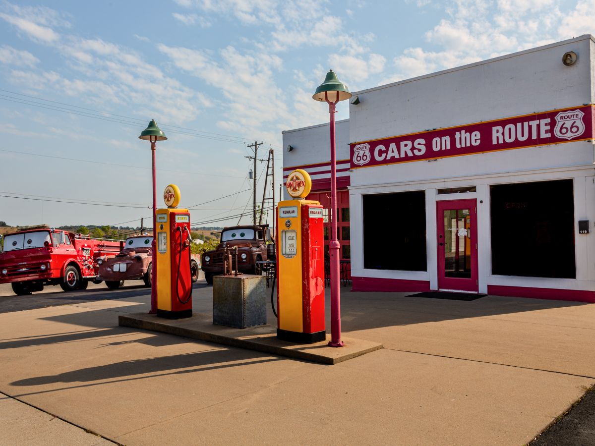 Gas station with vintage pumps on Route 66, featuring cartoon-style vehicles resembling a tow truck and firetruck. The building has "Cars on the Route" signage and a retro aesthetic.