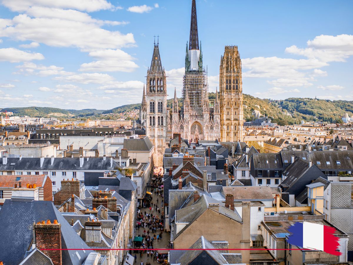 A view of Rouen, France, showcasing Gothic architecture with two prominent towers, surrounded by buildings under a blue sky with clouds.