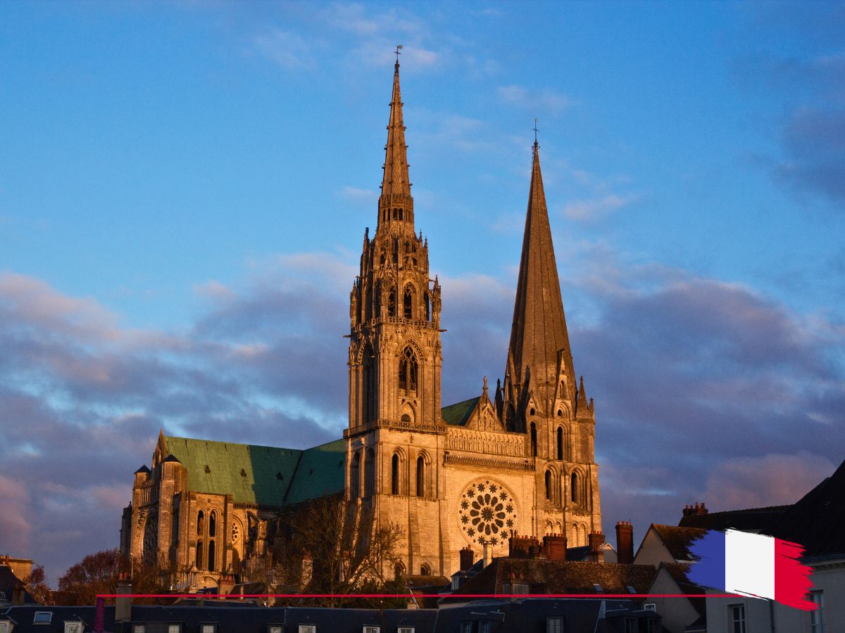 Historic cathedral with two distinct spires in a sunset glow, framed by rooftops, under a partly cloudy sky. A small French flag is displayed in the corner.
