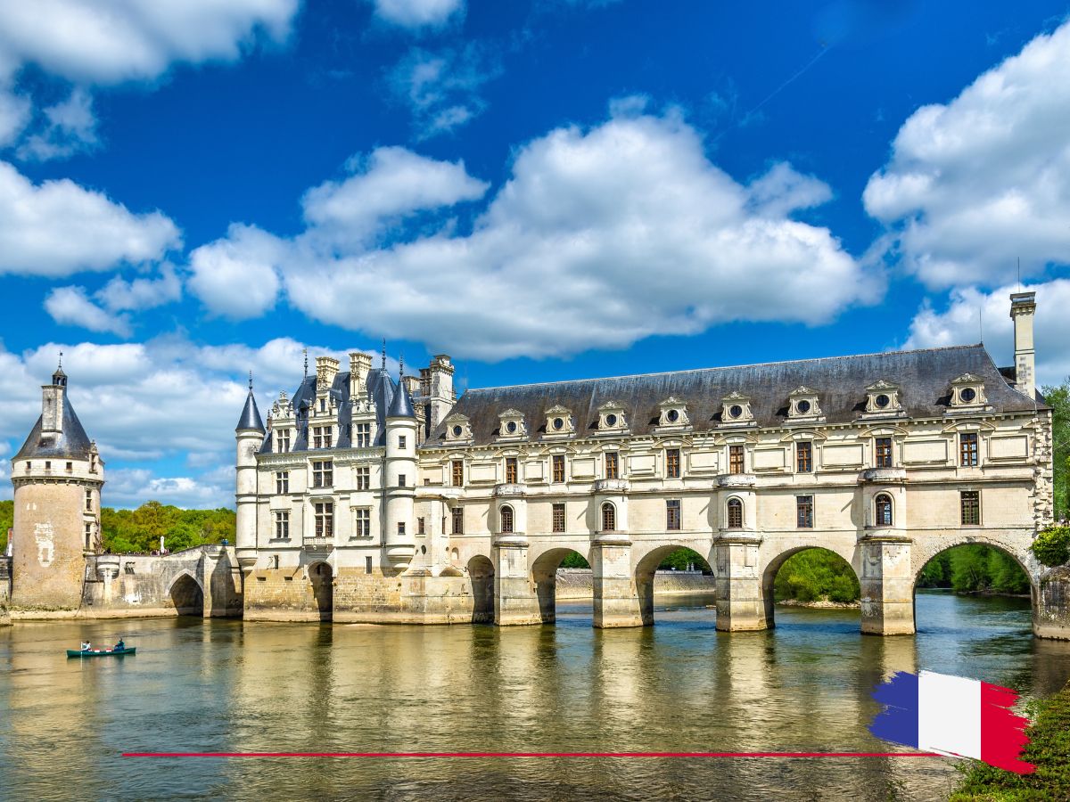 A historic chateau spans a river under a blue sky with clouds. A small boat is on the water. A French flag is in the bottom right corner.