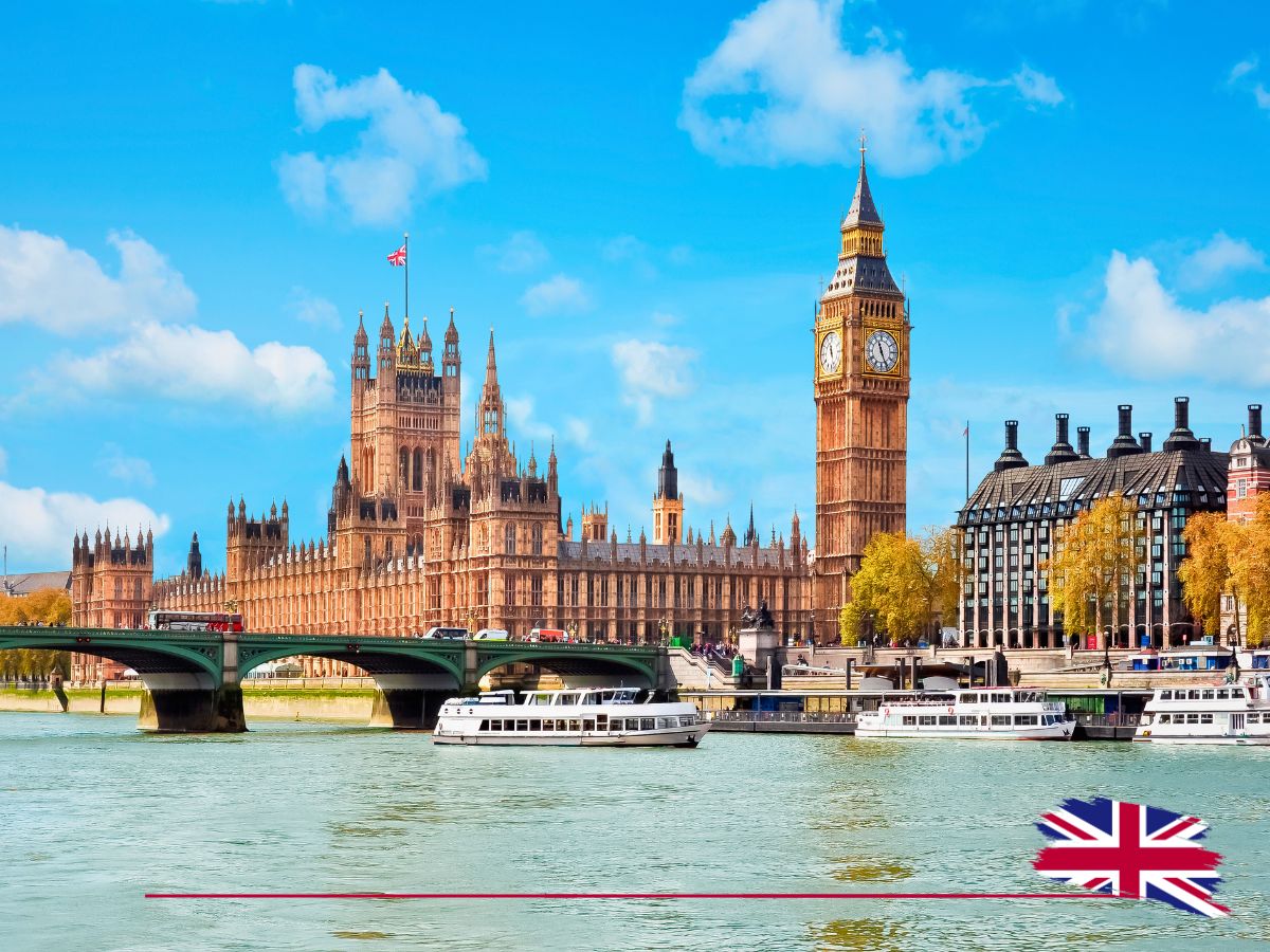 The Houses of Parliament and Big Ben with boats on the River Thames on a clear day, featuring a small British flag in the corner.