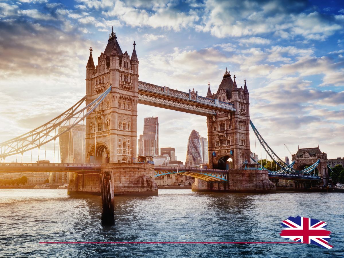 Tower Bridge in London over the Thames River at sunset, with modern city buildings in the background and a British flag in the foreground.