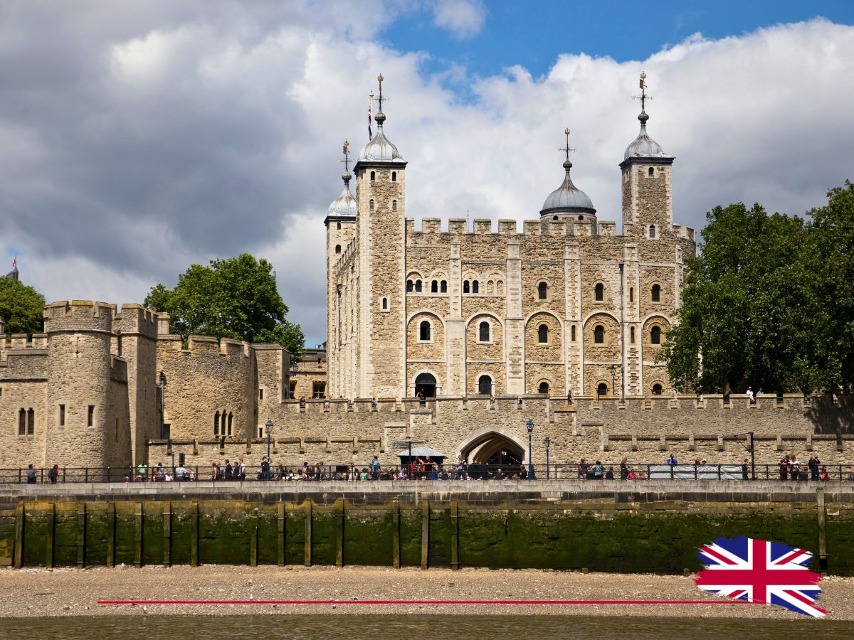 The Tower of London viewed from across the river, with people walking along the fortress walls. A Union Jack flag is in the foreground.